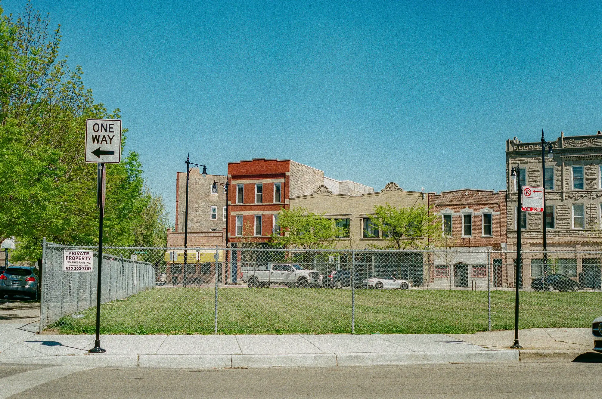 A grassed city lot behind a chain-link fence, mown short, with nothing built on it and row buildings on the far side